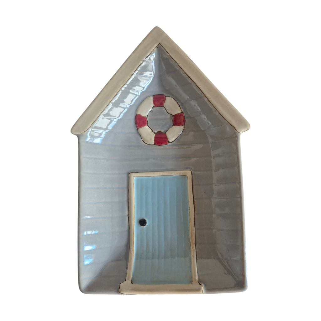 LaVida Beach House Plate — front view showing ceramic beach hut shaped plate with soft grey weatherboard exterior, white peaked roof, sky-blue door and red and white life ring wreath detail